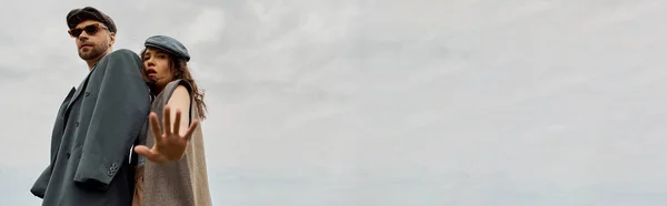 Vista de ángulo bajo de la mujer de moda en traje vintage mirando a la cámara mientras está de pie cerca de novio barbudo con chaqueta y gafas de sol con cielo nublado en el fondo, pancarta, escapada romántica - foto de stock