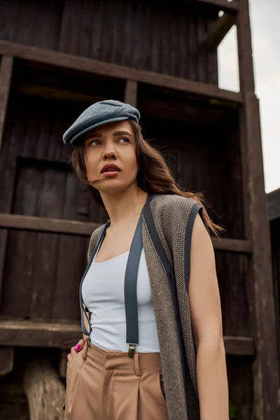 Portrait of fashionable brunette woman in newsboy cap, suspenders and vest holding hand in pocket of pants and looking away near rustic house in rural setting, vintage-inspired clothing — Stock Photo