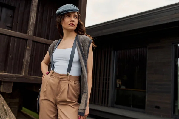 Fashionable brunette woman in vintage outfit, newsboy cap and suspenders holding hand in pocket of pants while standing near rustic house outdoors, vintage-inspired clothing — Stock Photo