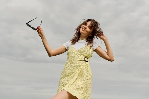 Low angle view of cheerful and stylish brunette woman in sundress looking at camera and holding sunglasses while standing with cloudy sky at background, summertime joy — Stock Photo
