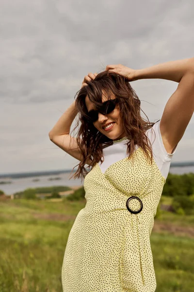 Portrait of joyful and stylish brunette woman in sundress and sunglasses touching head and standing with blurred scenic landscape and sky at background, summertime joy — Stock Photo
