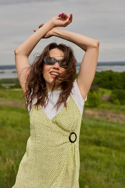 Retrato de mujer morena de moda y alegre en gafas de sol y vestido de sol posando mientras está de pie con el paisaje natural borroso y el cielo al fondo, alegría de verano - foto de stock