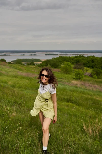 Mujer morena alegre y elegante en gafas de sol y vestido de pie en el prado verde con hierba y pasar tiempo con el paisaje borroso y el cielo en el fondo, alegría de verano - foto de stock