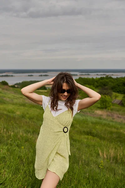 Mujer morena de moda y sonriente en gafas de sol y vestido elegante tocando la cabeza y de pie mientras pasa tiempo en el prado borroso borroso en el fondo, alegría de verano - foto de stock