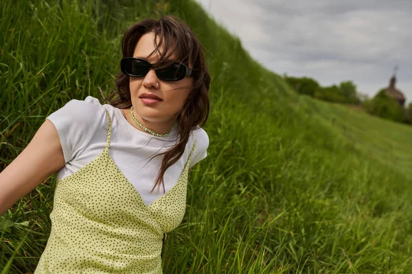 Portrait of trendy brunette woman in sunglasses and stylish sundress sitting on hill with blurred green grass and cloudy sky at background, natural landscape concept — Stock Photo