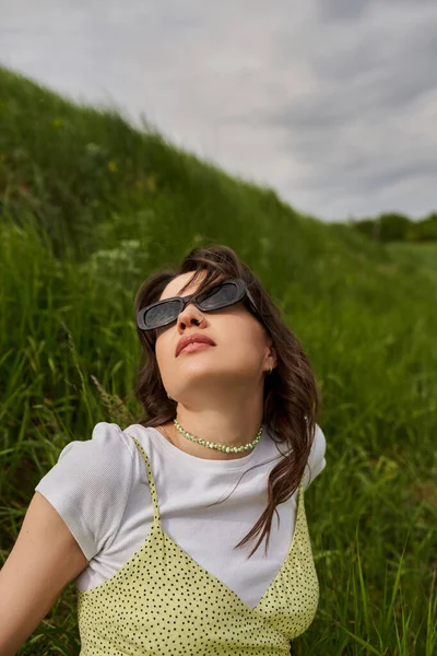 Портрет модной брюнетки в солнцезащитных очках и стильном солнечном платье сидя и расслабляясь на размытом травянистом холме с размытым ландшафтом и небом на заднем плане, природный ландшафт — стоковое фото