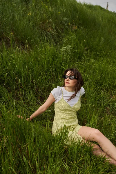 Mulher morena elegante em óculos de sol e vestido de sol relaxante e passar o tempo sentado na colina com grama verde no fundo, paisagem natural e conceito de espírito livre — Fotografia de Stock