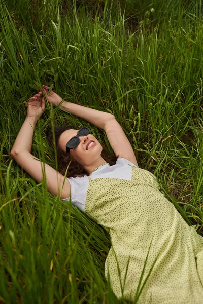 Vue de dessus de femme brune gaie et élégante en lunettes de soleil et robe de soleil couché et relaxant sur pelouse herbeuse en été, paysage naturel et relaxant dans le concept de la nature, paysage rural — Photo de stock
