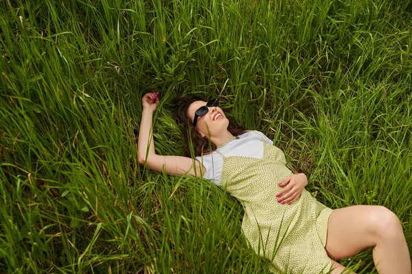 Blick aus der Vogelperspektive auf positive und stilvolle brünette Frau mit Sonnenbrille und Sonnenbrille liegend und entspannend auf Wiese mit grünem Gras, Naturlandschaft und entspannendem Naturkonzept — Stockfoto