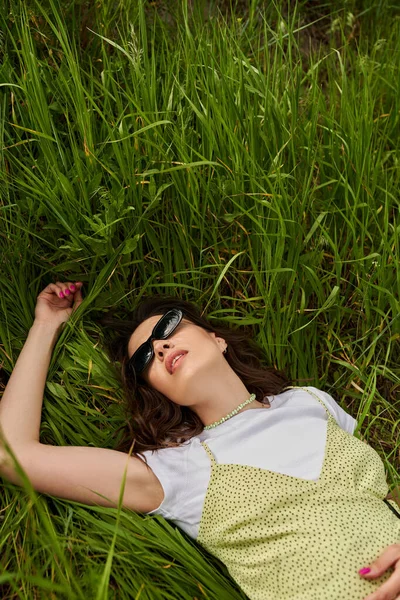 Vue du dessus de la femme brune à la mode en lunettes de soleil et robe de soleil couché sur l'herbe verte sur le terrain en été, paysage naturel et relaxant dans le concept de la nature, paysage rural — Photo de stock