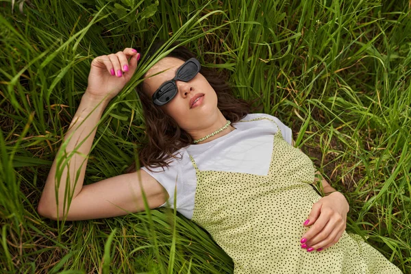 Vista superior de la mujer morena de moda en gafas de sol y vestido de sol relajante sobre hierba verde en el prado en verano, paisaje natural y relajante en concepto de naturaleza, paisaje rural - foto de stock