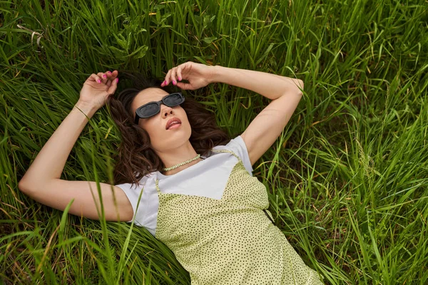 Vista superior de la mujer morena con estilo en gafas de sol y vestido de sol acostado en el prado herboso y relajante, retiro tranquilo y relajante en el concepto de la naturaleza, paisaje rural - foto de stock