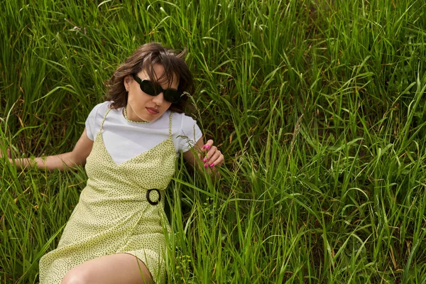 High angle view of fashionable brunette woman in sunglasses and sundress touching green grass while relaxing, peaceful retreat and relaxing in nature concept, rural landscape — Stock Photo