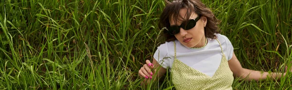 Hochwinkelblick der stilvollen brünetten Frau mit Sonnenbrille und Sonnenbrille auf der Wiese liegend und das grüne Gras berührend, friedlicher Rückzug und Entspannung in der Natur Konzept, Banner, ländliche Landschaft — Stockfoto