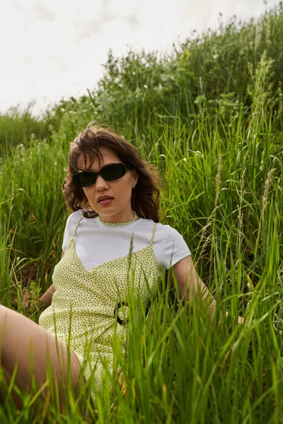 Portrait of stylish brunette woman in sunglasses and sundress relaxing on hill with green grass at summer, peaceful retreat and relaxing in nature concept, rural landscape — Stock Photo
