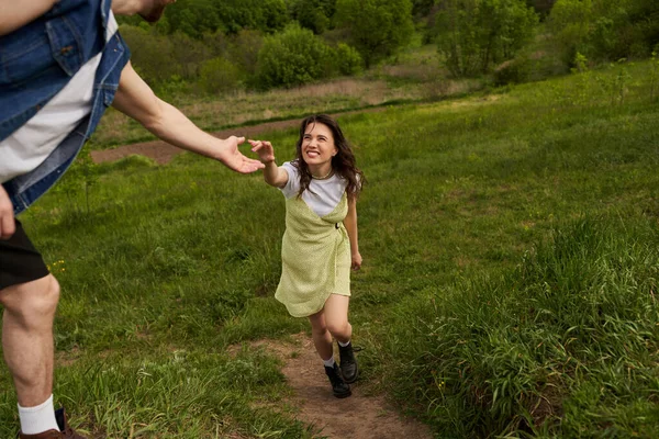 Cheerful brunette woman in stylish sundress and boots holding hand of boyfriend while walking on grassy hill at summer, rural tranquility and relaxing in nature concept — Stock Photo