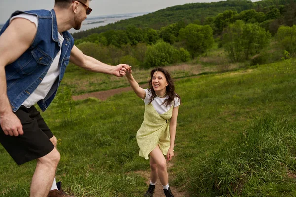 Stylish bearded man in sunglasses holding hand of cheerful brunette girlfriend in boots and sundress while walking on green hill, couple in love enjoying nature and relaxing concept — Stock Photo