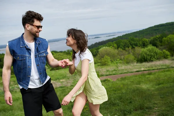 Smiling and stylish man in sunglasses holding hand of cheerful girlfriend in sundress while spending time and standing together with scenic landscape at background, couple in love enjoying nature — Stock Photo