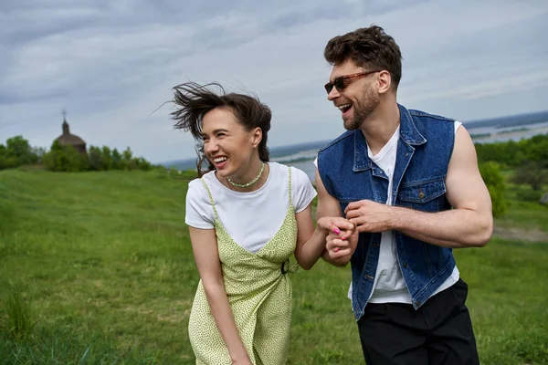Cheerful bearded man in sunglasses and denim vest holding hand of brunette girlfriend in sundress while walking on grassy field, couple in love enjoying nature and relaxing concept — Stock Photo