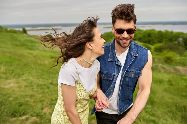 Cheerful brunette woman in sundress holding hand of stylish boyfriend in sunglasses and denim vest and walking on blurred grassy field, couple in love enjoying nature, tranquility — Stock Photo
