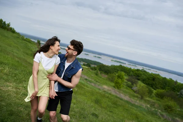 Couple romantique joyeux dans des tenues d'été élégantes s'amuser et se regarder tout en marchant sur une colline herbeuse floue avec ciel en arrière-plan, couple amoureux profiter de la nature, tranquillité — Photo de stock