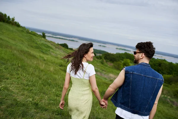 Vista lateral de casal romântico sorridente e na moda em roupas de verão de mãos dadas enquanto caminha em colinas gramadas com céu nublado no fundo, casal apaixonado apreciando a natureza, tranquilidade — Fotografia de Stock
