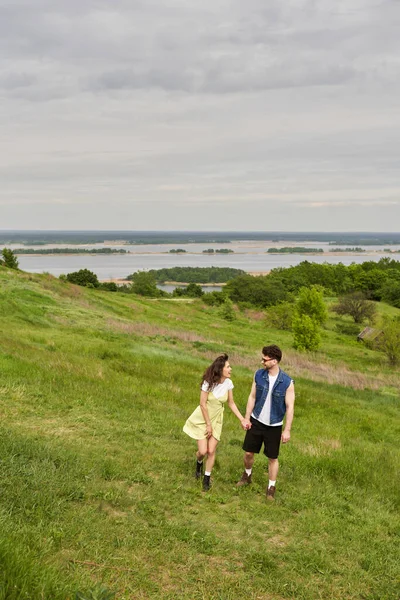 Cheerful brunette woman in sundress and boots talking to stylish boyfriend in sunglasses and denim vest and walking on hill with grass together, couple in love enjoying nature, tranquility — Stock Photo