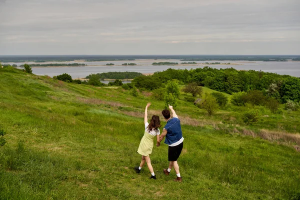 Back view of man in stylish denim vest pointing with finger and holding hand of girlfriend in sundress and standing on grassy field together, couple in love enjoying nature, tranquility — Stock Photo