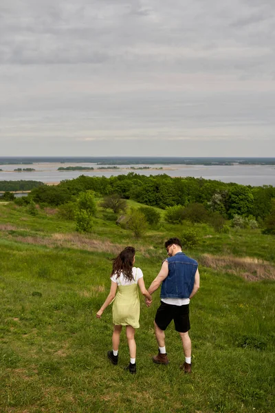 Full length of stylish brunette romantic couple in summer outfits holding hands while relaxing and walking on green hill with scenic landscape at background, countryside adventure and love story — Stock Photo
