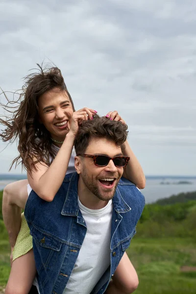 Cheerful brunette woman piggybacking on stylish boyfriend in sunglasses and denim vest while standing with blurred landscape at background, countryside adventure and love story, tranquility — Stock Photo