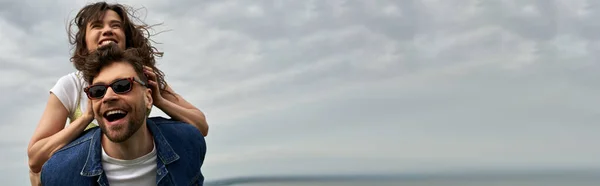 Positive brünette Freundin im Sommeroutfit huckepack auf stylischem Freund in Sonnenbrille und Jeansweste im Stehen mit wolkenverhangenem Himmel im Hintergrund, Abenteuer auf dem Land und Liebesgeschichte, Banner — Stockfoto