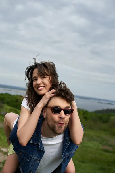 Cheerful brunette woman touching hair of excited and stylish boyfriend in sunglasses while piggybacking and having fun with scenic nature at background, countryside adventure and love story — Stock Photo
