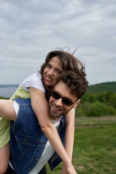 Cheerful brunette woman in sundress hugging stylish boyfriend in sunglasses and denim vest while piggybacking and looking at camera with landscape at background, countryside adventure and love story — Stock Photo