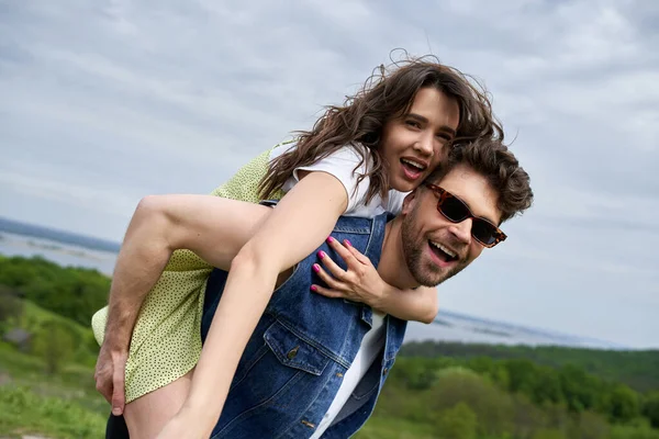 Excited stylish woman in sundress piggybacking on cheerful boyfriend in denim vest and sunglasses while spending time on nature, countryside adventure and love story, tranquility — Stock Photo