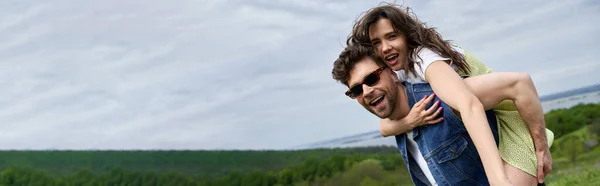 Fröhliche brünette Frau im schicken Outfit huckepack auf bärtigen Freund in Sonnenbrille und Jeansweste und Zeit für Natur, Abenteuer auf dem Land und Liebesgeschichte, Banner — Stockfoto