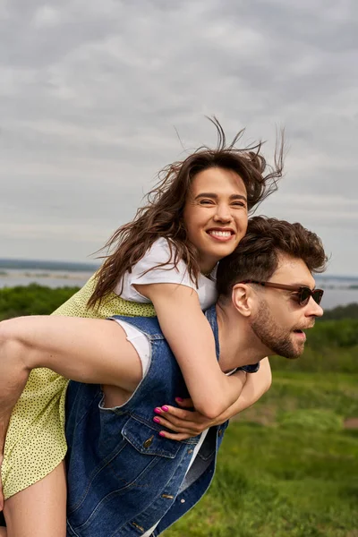 Joyful brunette woman in sundress hugging bearded boyfriend in sunglasses and denim vest and having fun on nature at background, countryside adventure and love story, tranquility — Stock Photo