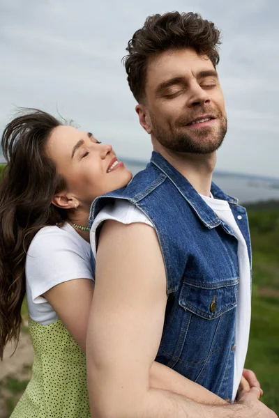 Portrait of brunette woman in sundress smiling and hugging bearded boyfriend in denim vest and closing eyes while standing with nature at background, love story and countryside adventure — Stock Photo