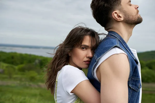 Portrait of brunette woman looking at camera while embracing bearded boyfriend in stylish denim vest and spending time with tranquility landscape at background, love story and countryside adventure — Stock Photo