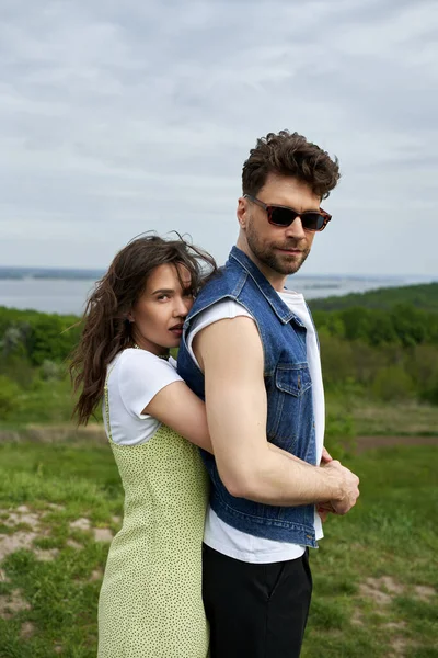 Stylish woman in sundress embracing bearded boyfriend in sunglasses and denim vest and looking at camera while standing with landscape and sky at background, love story and countryside adventure — Stock Photo
