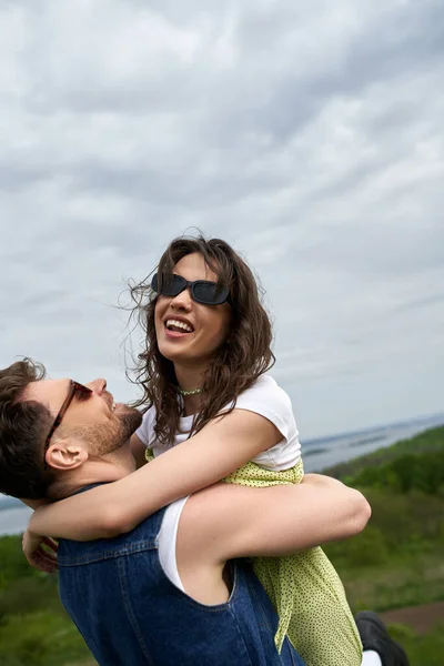 Cheerful bearded man in denim vest ad sunglasses hugging stylish brunette girlfriend in sundress and spending time in rural landscape at background, love story and countryside adventure — Stock Photo