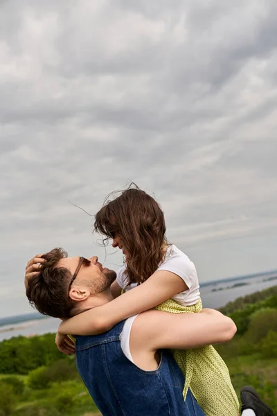 Side view of smiling and trendy man in sunglasses and denim vest hugging and lifting brunette girlfriend in sundress while spending time in rural setting, love story and countryside adventure — Stock Photo