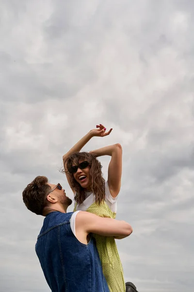 Homme brune gai en gilet de denim élégant tenant petite amie dans des lunettes de soleil et robe de soleil et s'amuser avec ciel nuageux en arrière-plan à l'été, histoire d'amour et aventure à la campagne — Photo de stock