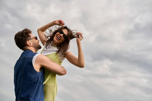 Homem barbudo alegre em colete jeans levantando namorada morena em roupa de verão e óculos de sol e de pé com céu nublado no fundo, história de amor e aventura no campo, tranquilidade — Fotografia de Stock