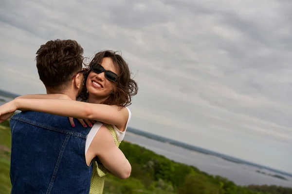 Mulher morena sorridente em roupa de verão e óculos de sol abraçando namorado elegante em colete jeans com paisagem rural e céu nublado no fundo, história de amor e aventura no campo — Fotografia de Stock