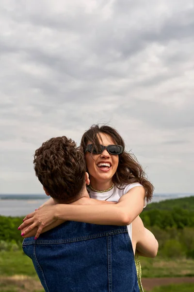 Morena alegre namorada em óculos de sol e roupa de verão abraçando e se divertindo com namorado em colete jeans enquanto está em ambiente rural, história de amor e aventura no campo — Fotografia de Stock