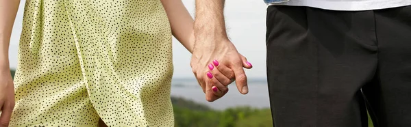 Cropped view of stylish couple in summer clothes holding hands while standing together with blurred landscape at background, countryside wanderlust and love concept, banner, tranquility — Stock Photo