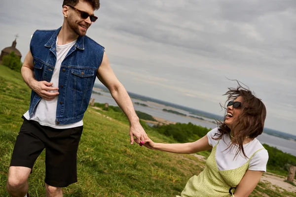 Hombre barbudo sonriente en gafas de sol y chaleco vaquero elegante cogido de la mano de una novia alegre en vestido de noche y pasar tiempo en la colina cubierta de hierba en el fondo, la lujuria rural y el concepto de amor - foto de stock