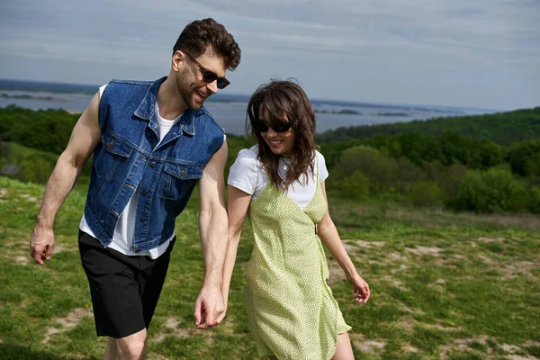 Positive love couple in sunglasses and stylish summer outfits holding hands while spending time on blurred landscape at background, countryside wanderlust and love concept, tranquility — Stock Photo