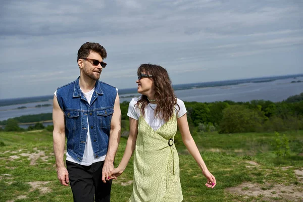 Cheerful brunette woman in sunglasses and stylish sundress holding hand and looking at boyfriend in denim vest and standing on grassy hill, countryside wanderlust and love concept, tranquility — Stock Photo