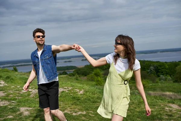 Smiling brunette man in sunglasses and denim vest holding hand of stylish girlfriend in sundress and spending time on grassy hill with cloudy sky at background, countryside wanderlust and love concept — Stock Photo